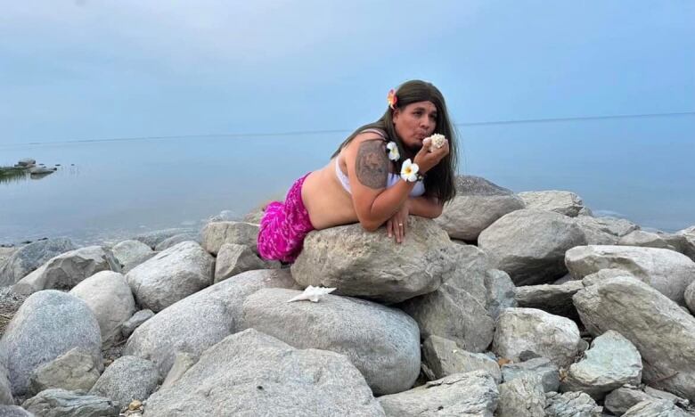 A man lies on a pile of rocks in a merman costume, with long flowing hair, a pink fin, a white bra and flowers in his hair and on his wrist. 