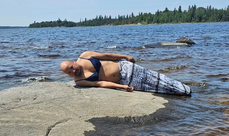 A man lies on a rock in a merman costume, with a blue bra and patterned fin.