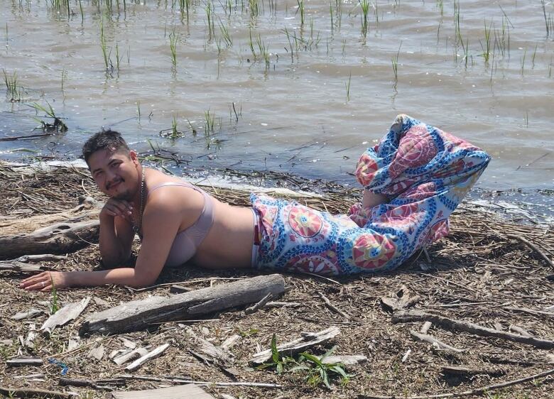 A man lies in a patterned merman costume next to a river.