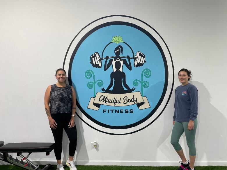 Two women posing in front of a sign in a gym that reads Mindful Body Fitness 