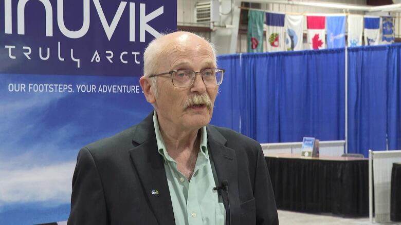 An elderly man stands in a room in front of an Inuvik sign.