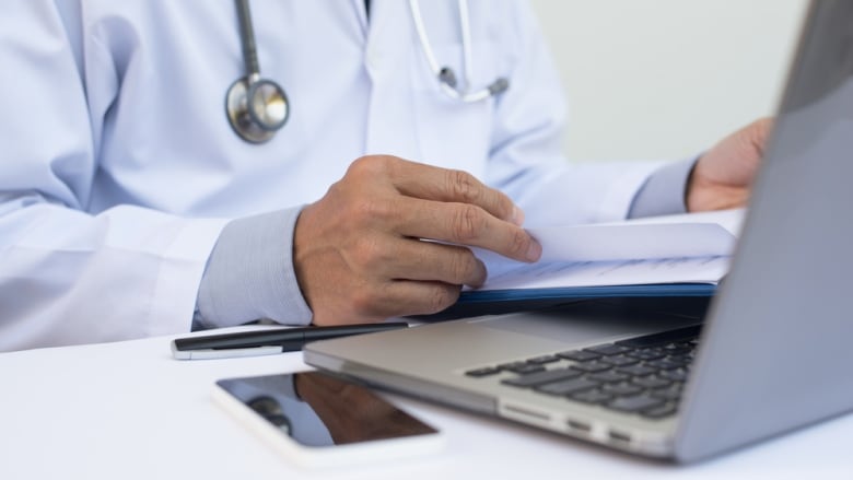 Doctor reading patient health record chart and working on laptop computer on white desk in hospital with copy space, electronic health records system EHRs, teleconference or telemedicine concept.