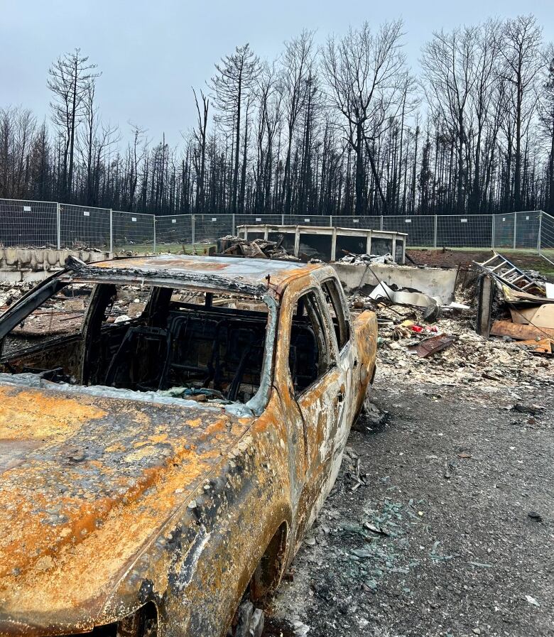 A burnt-out car is shown in front of a burnt property surrounded by white fencing. 