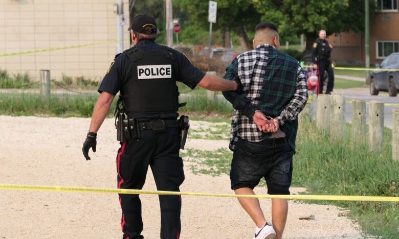 A police officer is seen from the back, leading away a man who is wearing handcuffs and wearing shorts.