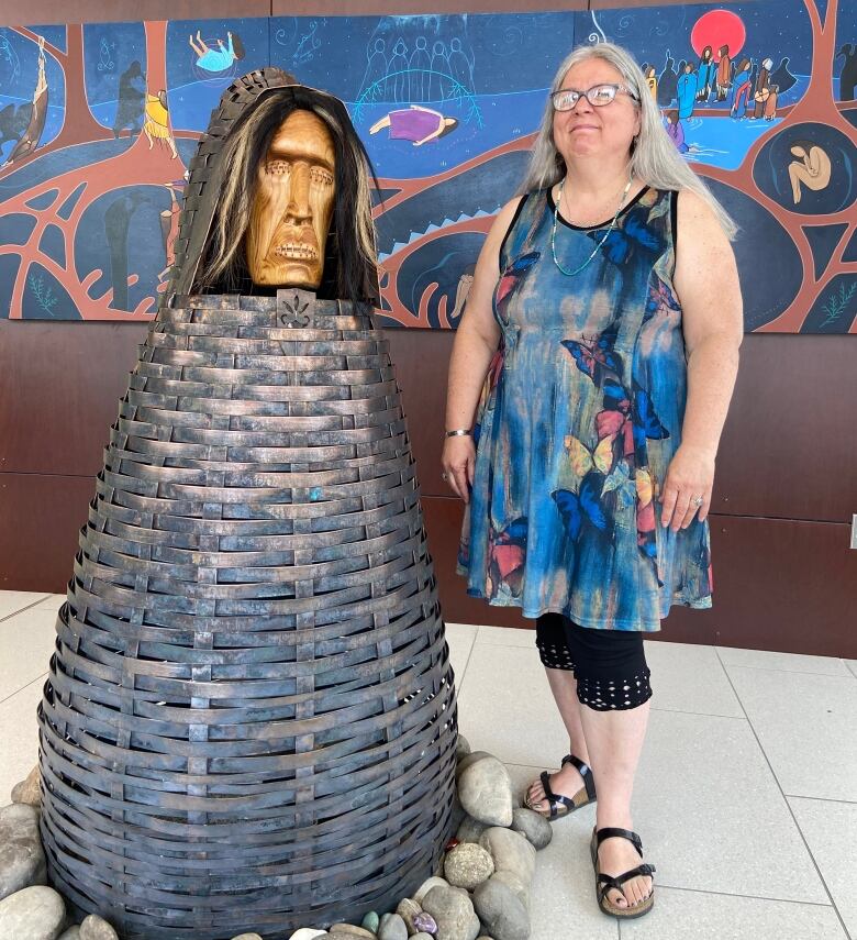 Women with gray hair and glasses stands next to statue with eyes and mouth sewn shut.