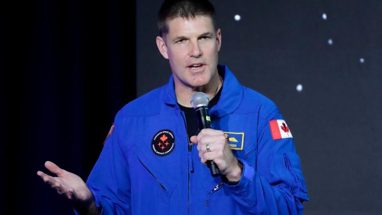 A man wearing a blue uniform with patches on it speaks holding a microphone.