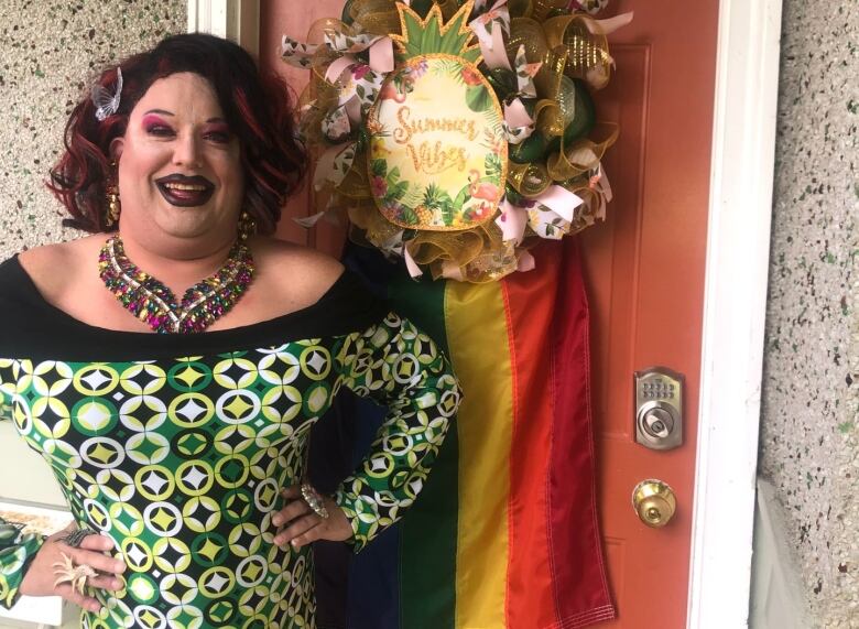 A person in a patterned green and yellow dress, a multi-coloured bejewelled necklace, purple drag makeup and a deep red bob haircut, poses in front of a door that has a floral sign saying 'Summer Vibes' and a LGBTQ flag hanging on it.