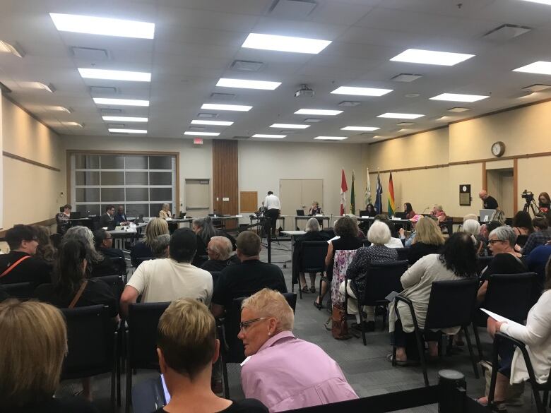 A school board office shows people sitting in chairs, with flags at the front of the room.