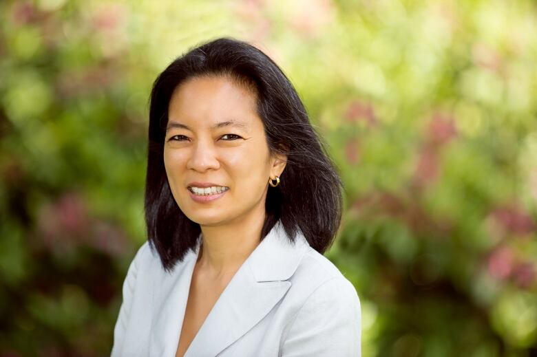 A woman with dark hair and light blazer stands in front of leafy, blurred background.