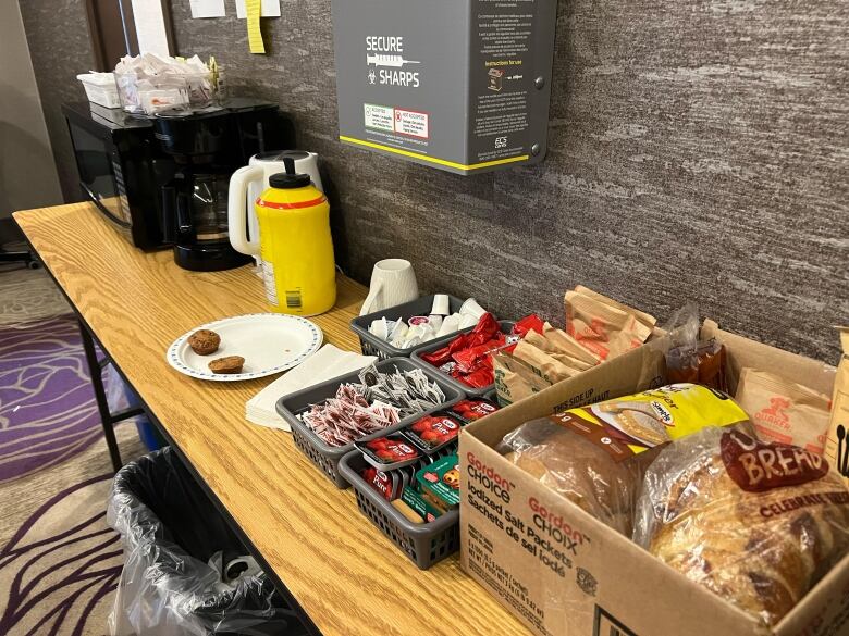 A table is shown with a basket of bread, jam and peanut butter packets, oatmeal packets, condiments, a coffee machine and a microwave atop.