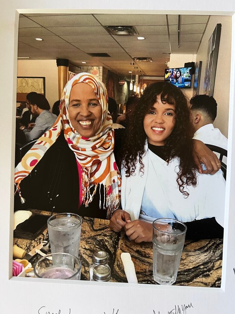 Two woman sit with their arms around each other, smiling into the camera at a local restaurant. 