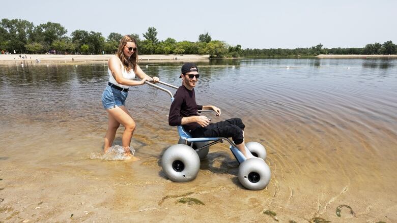 A woman with long blonde hair pushes a man in a backward baseball cap and sunglasses in a specialized wheelchair in shallow lake water.