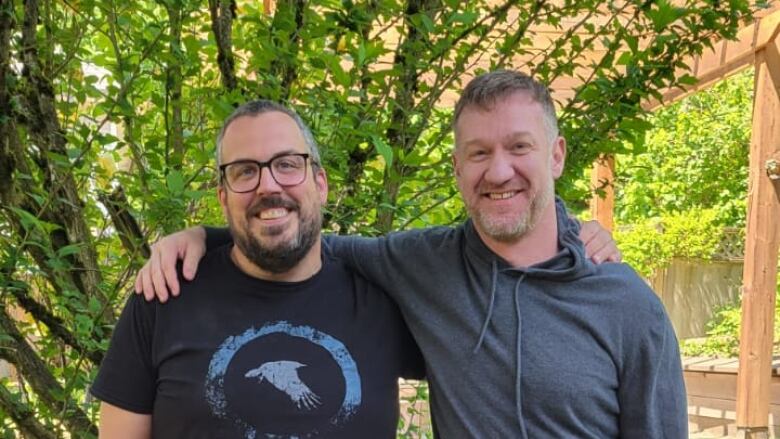 Two men stand in a backyard with their arms around each other smiling.