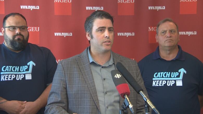 A man stands behind a podium, while he's flanked behind him by some supporters.