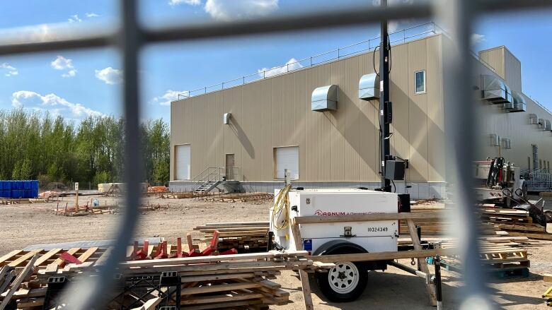 A large industrial building is seen behind a fence with construction material in the foreground.