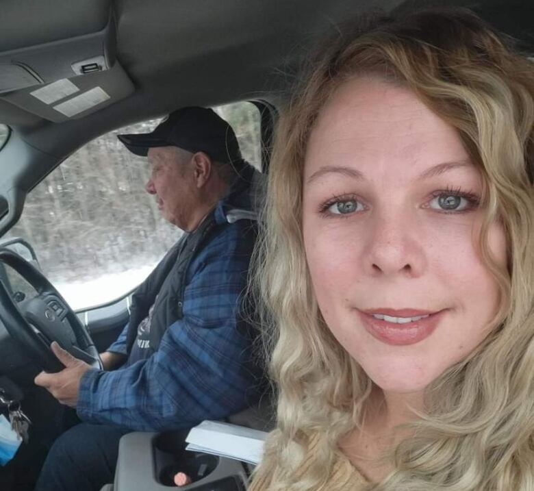 A smiling woman is seen in the passenger seat of a vehicle with a man in the background, driving. 