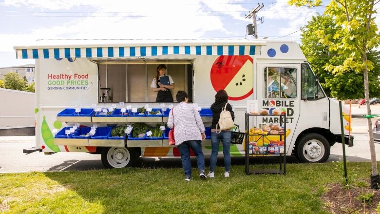 A white truck with a blue and white striped awning doubles as a food market. There are paintings of produce on the side like apples, peas carrots, bananas and pears. Someone is inside the vehicle holding a clipboard and two customers are shopping outside. 