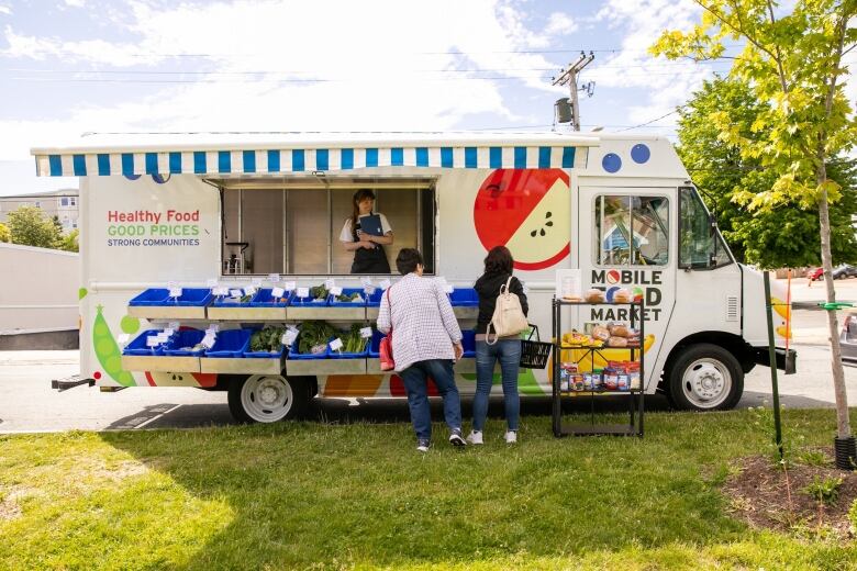 A white truck with a blue and white striped awning doubles as a food market. There are paintings of produce on the side like apples, peas carrots, bananas and pears. Someone is inside the vehicle holding a clipboard and two customers are shopping outside. 