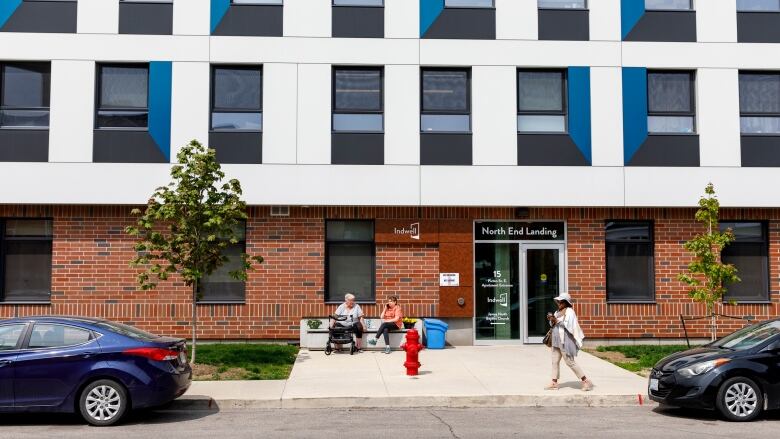 Two people site and chat on a bench in front of a building that's brick on the first floor and white above as a woman walks past.