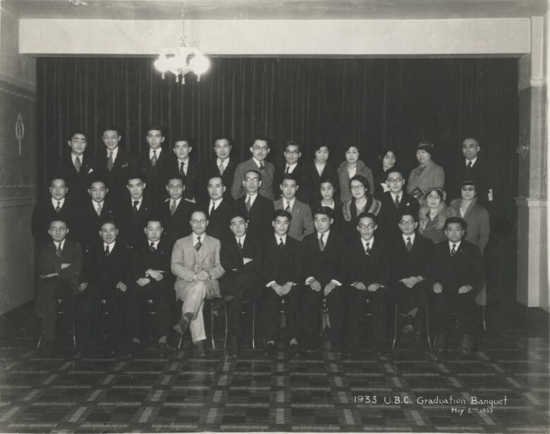 A group of people in suits sit and stand in rows inside a room with a curtain in the background.