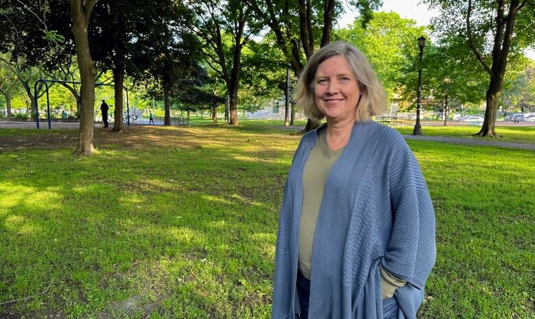 A woman wearing a cardigan stands outside on a sunny day, amid trees.