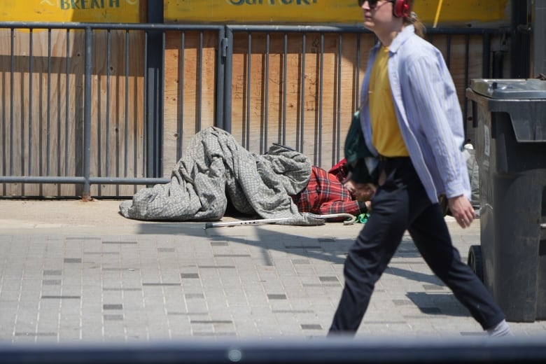 A man in a sleeping bag sleeps in the street as someone walks by seemingly unaware