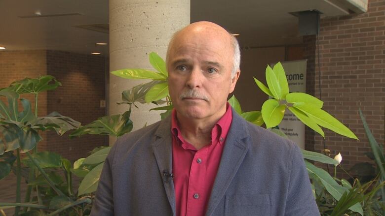 A bald man stands in front of some plants in a lobby area.