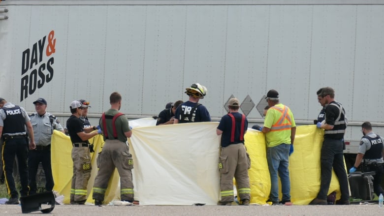 A group of police and firefighters at the scene of a highway crash hold up yellow tarps near a semi-trailer truck.