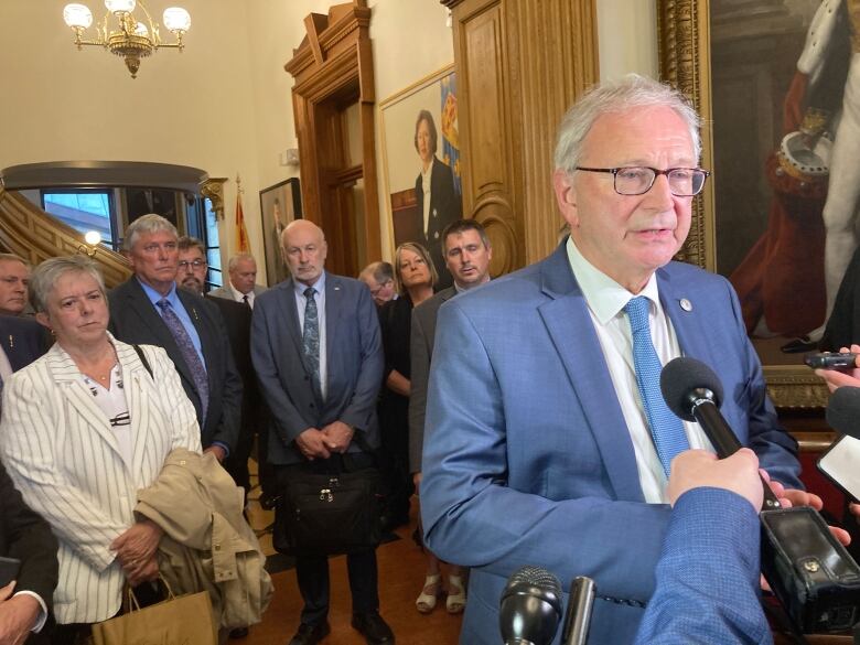 A man with grey hair and glasses, wearing a blue suit, white collared shirt and blue tie, speaks into several reporters' microphones as a number of other people behind him look on.