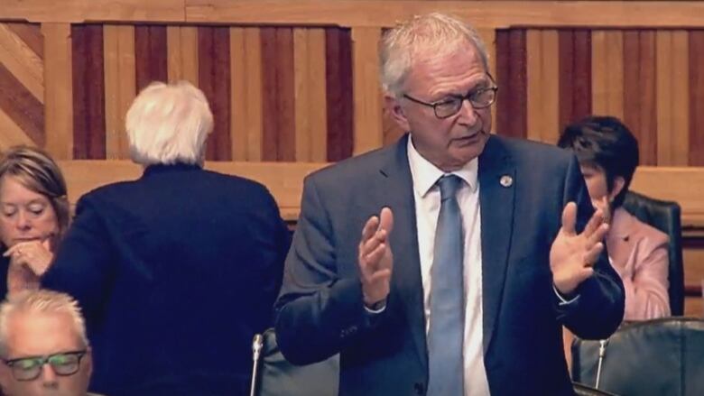 A man speaking at a podium surrounding by people sitting down. A woman on the left is standing with her back to the speaker.