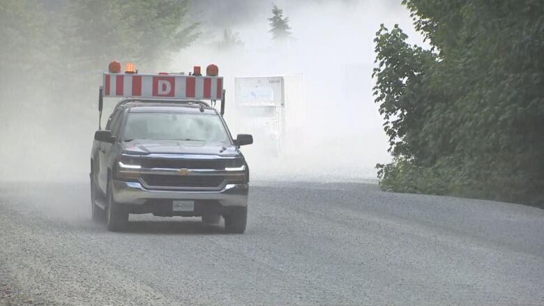 A truck with a large warning sign and lights on it drives along a dusty gravel road with trees all around.