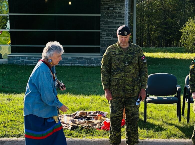 A woman stands outside gesturing toward the ground. To her right is a man dressed in green camouflage clothing. 