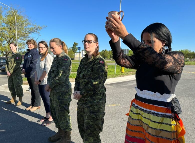 A woman wearing a colourful skirt holds up cup. People wearing green camouflage line up next to her.