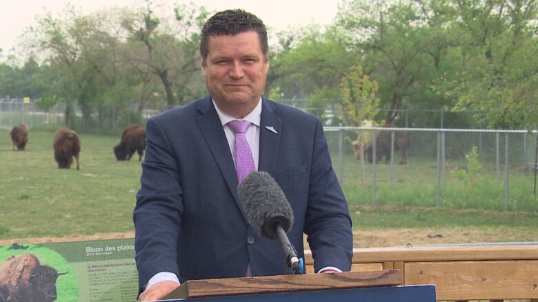 A man stands at a podium, in front of an enclosure of bisons.