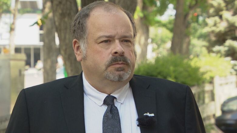 A man wearing a black tie and suit jacket stands in front of trees.