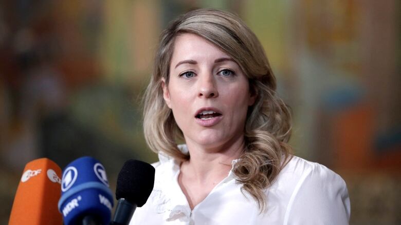 A woman in a white shirt speaks near three microphones.