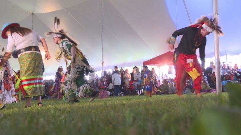 Two First Nations powwow dancers wearing traditional garments dance to the left of the frame, and to the far right, another First Nations dancer faces to the right of the frame while dancing. The dancers are under a large white tent, and a crowd of people watches in the background.