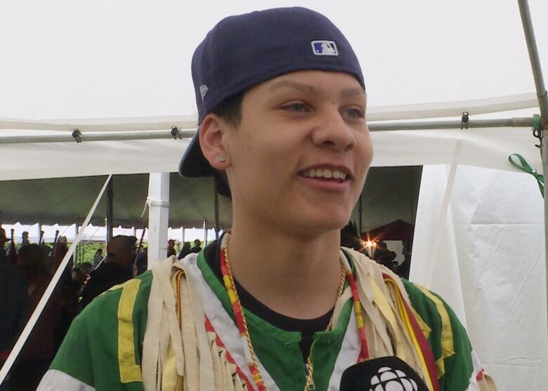 A young man in a backwards baseball cap smiles at the camera. He's wearing a green shirt with a multi-coloured traditional garment. He's standing in front of a large white event tent.