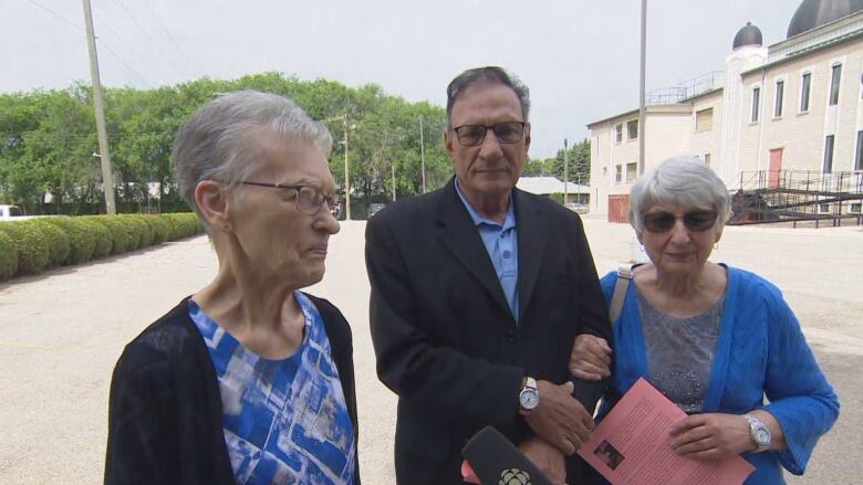 Three elderly people - two women and a man - stand in a parking lot behind a church. The woman on the left has short grey hair and is wearing glasses, and a black shawl on top of a blue patterned shirt. The man in the middle has short charcoal hair, glasses, a watch on his right wrist and is wearing a black dress coat over a blue buttoned shirt. The woman on the right has her right arm intertwined the mans left. She white hair, sunglasses, a watch on her left wrist and is wearing a blue sweater over a grey shirt. She is holding a pink sheet of paper.