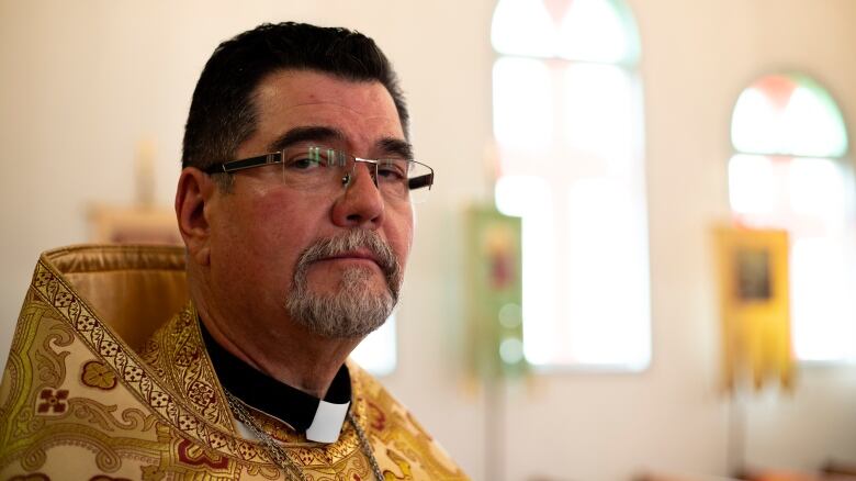 A white man, with short black hair that is greying near the sides, a grey goatee and glasses is standing inside a church. He is wearing a patterned cloak of gold, with dashes of red.