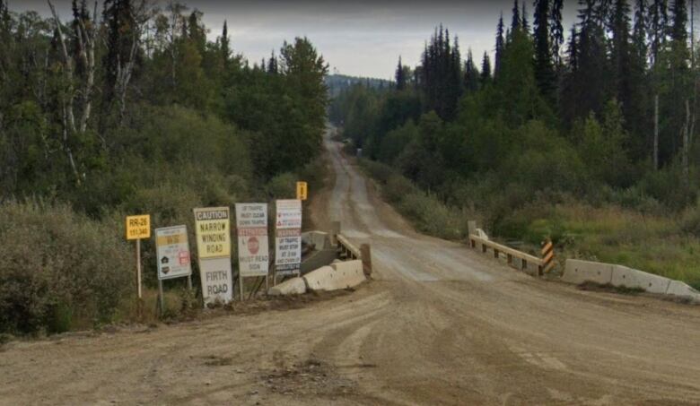 Signs are displayed at the entrance of a gravel road through a forested area, with warnings such as 