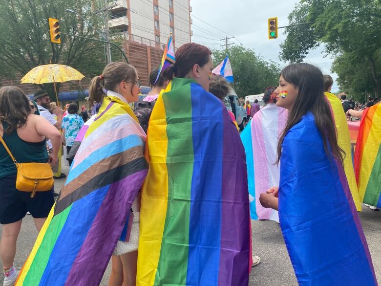 People are gathered for the pride parade. In the near view there are three people with a large rainbow flag.