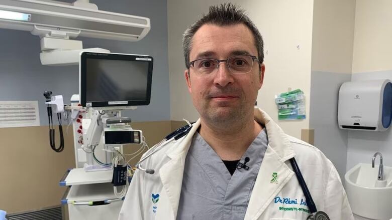 A man with short dark, greying hair, wearing glasses, medical scrubs, a white doctor's coat and stethoscope, stands in front of medical equipment.