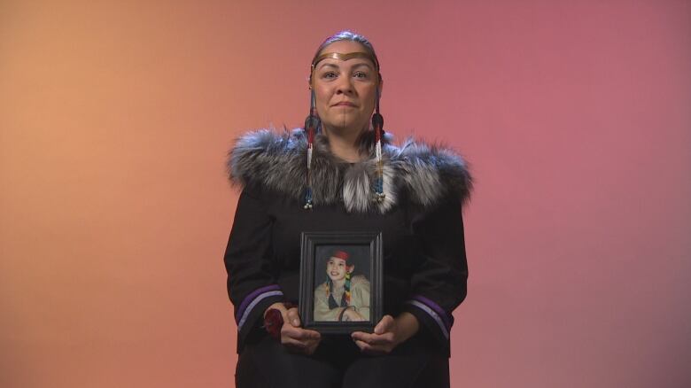 A woman with long dark hair, wearing a parka and headdress, holds a photo of a teenaged girl in her hands.