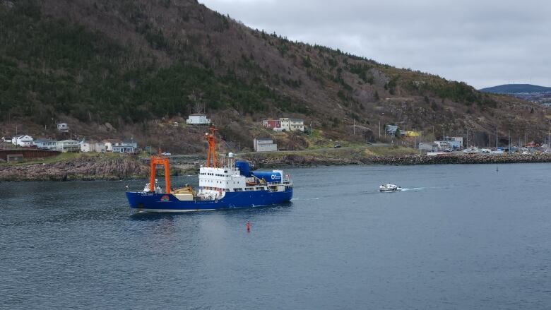 A large blue shipping vessel sails out to sea. A smaller, white submersible vehicle is towed behind.