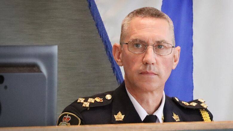 A man with short hair and glasses is in front of a Nova Scotia flag wearing a dark blue uniform with badges and pins on it.