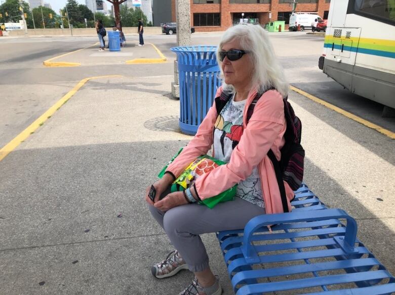 A woman with white hair wearing sunglasses, a white t-shirt, a pink jacket and grey pants sitting on a bench at a bus stop