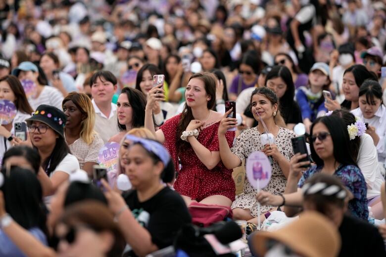 A crowd of people, mostly young women, sit on the ground at a large park.