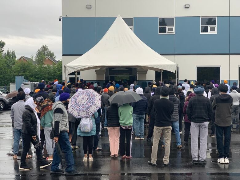 Hundreds of people gather outside a Sikh temple in rainy weather with a white tent-like awning over the entrance.