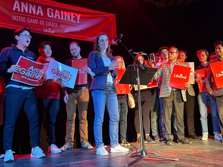 A group of people stand on stage with Anna Gainey posters and signs.